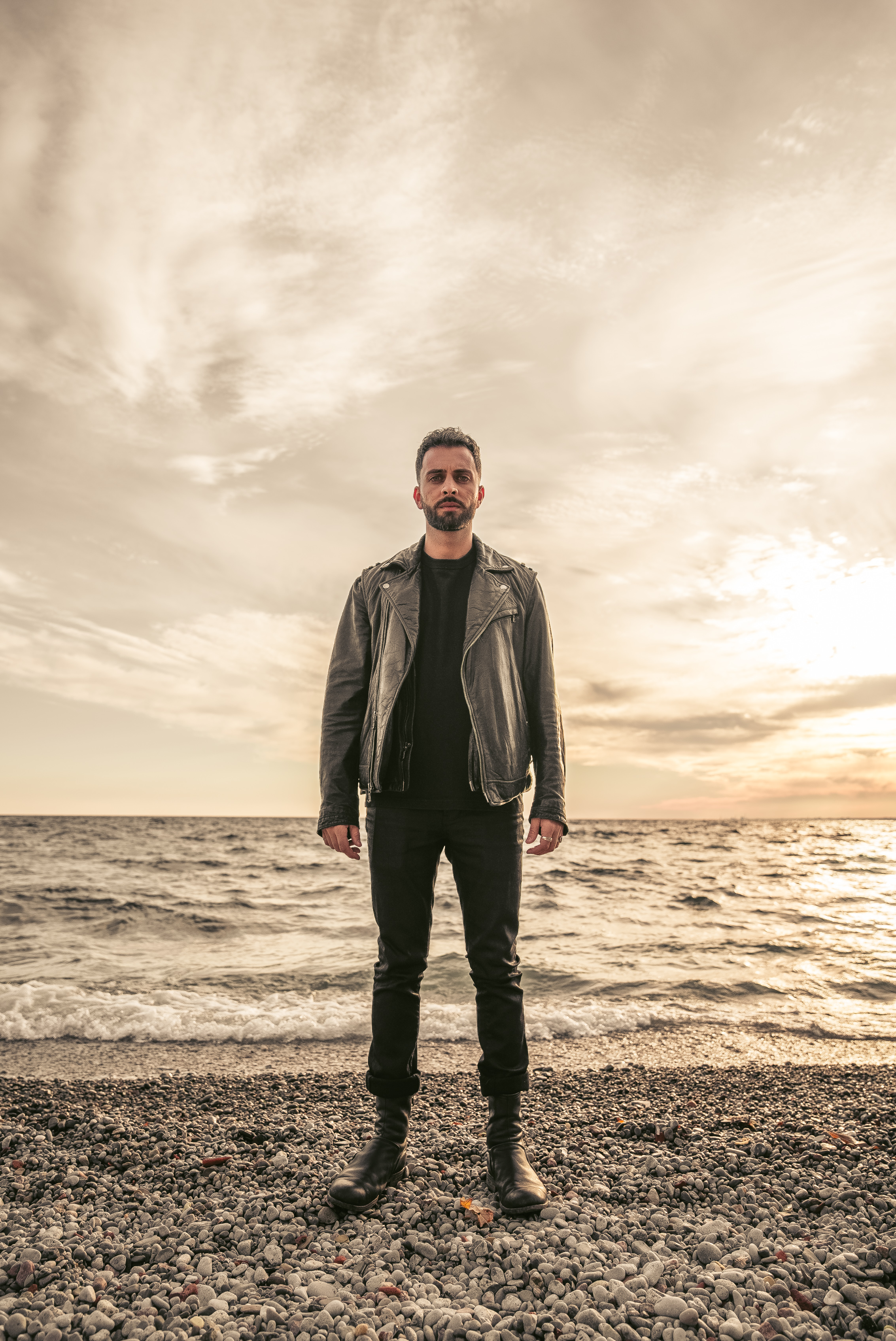 Man in leather jacket standing on rocky beach at golden hour with dramatic sky